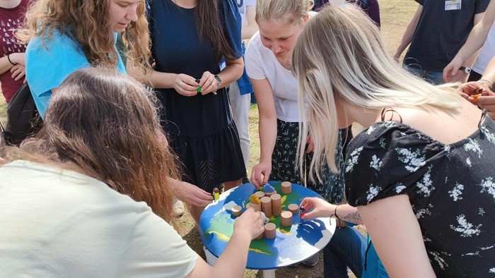 © Justus Braune A group of people is gathered around a small round table with a world map design, placing wooden blocks on its surface.