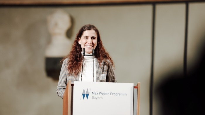 Eine Person hält eine Ansprache am Rednerpult mit der Aufschrift Max Weber Programm Bayern.