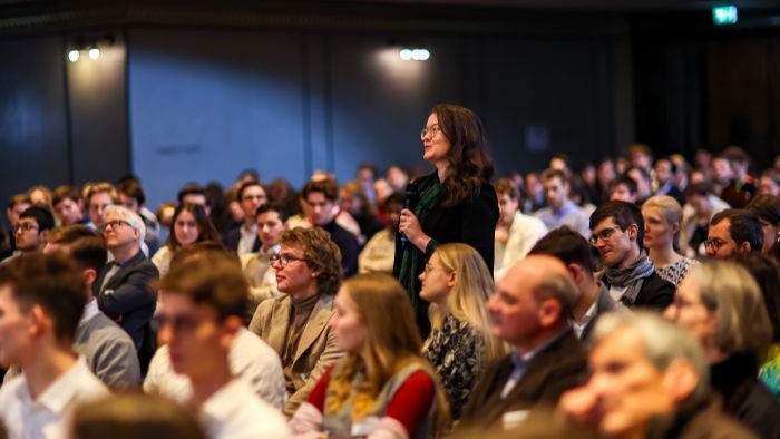 In einem voll besetzten Saal steht eine Person im Publikum auf und spricht in ein Mikrofon, während die übrigen Gäste aufmerksam zuhören.