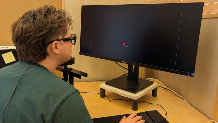 © Dario Neuhaus Eine Person sitzt an einem Schreibtisch vor einem großen Monitor, auf dem ein schwarzer Bildschirm mit einem roten Punkt und einem weißen Fadenkreuz angezeigt wird, während ein Kabel am Kopf der Person befestigt ist.