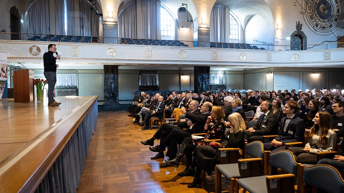 A man speaks on a stage to people sitting on chairs