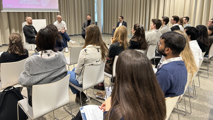 Vier Männer sitzen in einem hellen Raum in einem Halbkreis vor Publikum und diskutieren.