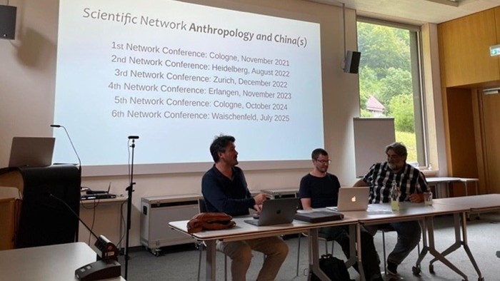 Drei Männer sitzen an einem Tisch vor einer Leinwand, die die Daten vergangener Konferenzen anzeigt.