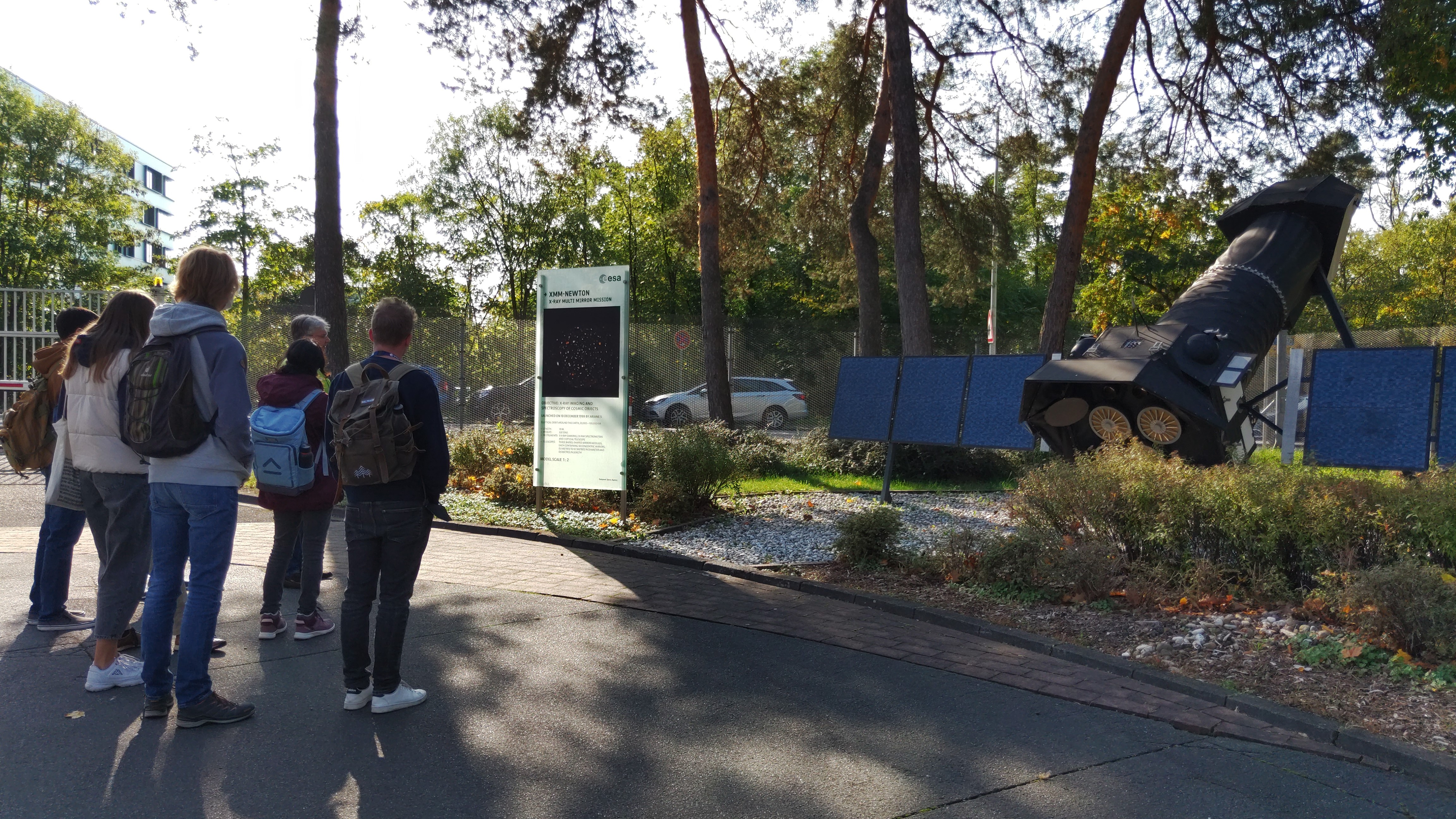 Eine Personengruppe betrachtet ein Satellitenmodell am Rande des ESOC-Geländes in Darmstadt mit Bäumen im Hintergrund.