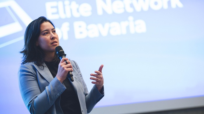 © Andreas Gebert A young woman in a light-colored blazer is standing on stage, speaking into a microphone.