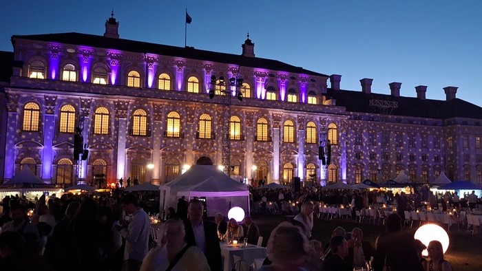 Menschen sitzen abends im beleuchteten Innenhof vor dem festlich angestrahlten Schloss Schleißheim.