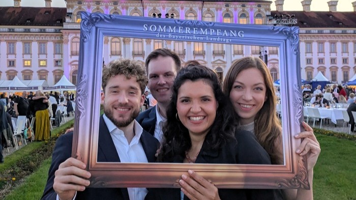Zwei Männer und zwei Frauen stehen vor einem Schloss und lächeln durch einen leeren Bilderrahmen in die Kamera.