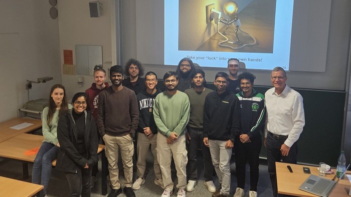 Eine Gruppe von Personen steht in einem Seminarraum vor einer Präsentationsfolie mit einer Glühbirnen-Grafik.