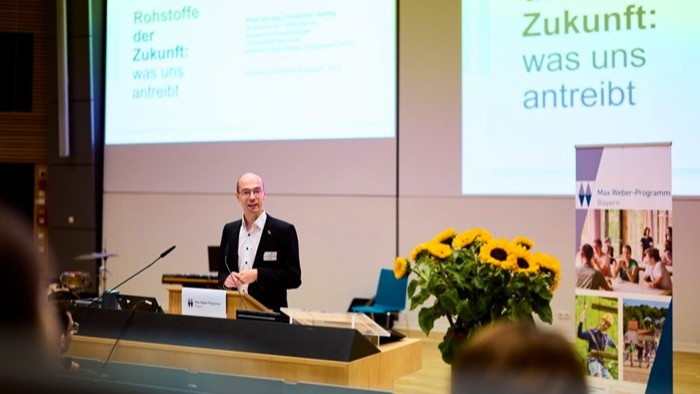 Ein Redner steht an einem Pult in einem Hörsaal und hält einen Vortrag zum Thema „Rohstoffe der Zukunft: was uns antreibt“. Neben dem Pult steht ein großer Strauß Sonnenblumen. Im Hintergrund sind eine Präsentationsfolie und ein Banner des Max Weber-Programms zu sehen.
