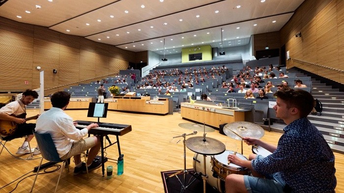 Eine Band mit Gitarre, Keyboard und Schlagzeug spielt auf der Bühne eines großen, modernen Hörsaals. Im Hintergrund sitzt das Publikum auf den Rängen und hört zu. 