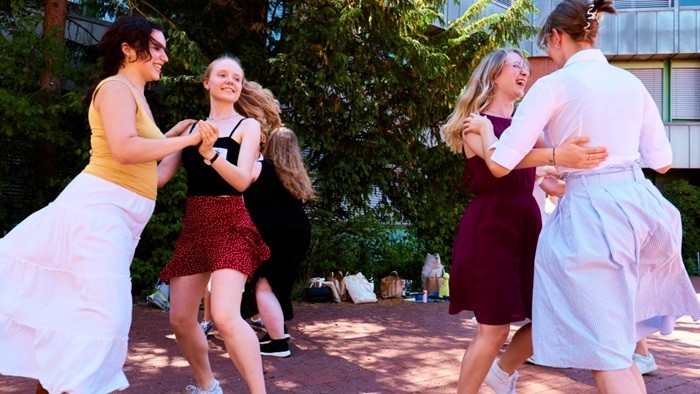 Mehrere junge Menschen tanzen paarweise im Freien auf einem sonnigen Platz. Sie tragen sommerliche Kleidung und bewegen sich fröhlich zur Musik. Im Hintergrund sind Bäume und ein Gebäude zu sehen.