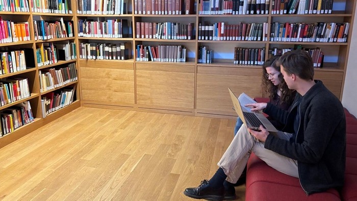 Eine junge Frau mit einem Buch und ein junger Mann mit einem Laptop sitzen auf einem Sofa und unterhalten sich.