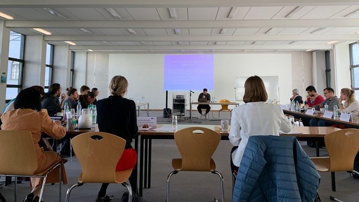 © Matthias Laufhütte, IDK Philologie Eine junger Mann spricht an einem Tisch vor einer Projektionsfläche.