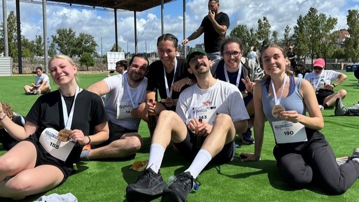 © Chiara Spear A group of people in athletic wear are sitting on the grass, beaming with joy, with medals hanging around their necks.