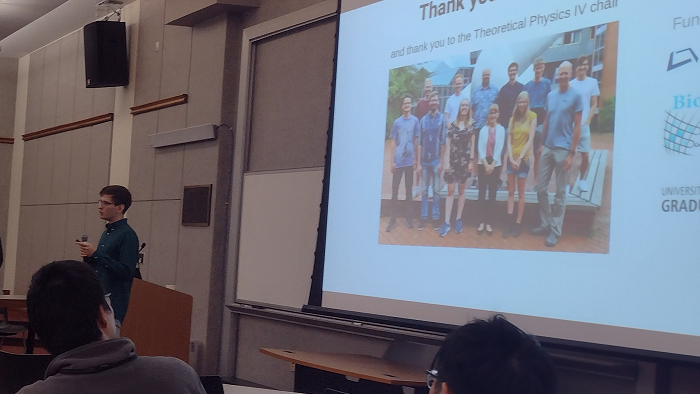 A young man is standing behind a lectern speaking in front of a screen. His acknowledgement slide is seen in the background.