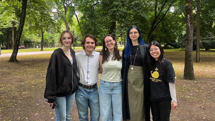 Fünf Menschen stehen nebeneinander in einem grünen Park mit vielen Bäumen, tragen lässige Kleidung und posieren für ein Foto.
