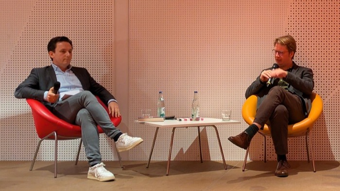 © Julian Werlitz Zwei Personen sitzen in bunten Sesseln auf einer Bühne und führen ein Gespräch, wobei ein Tisch mit Wasserflaschen und Gläsern zwischen ihnen steht.