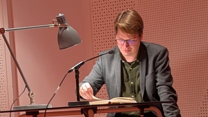 Eine Person steht an einem Pult mit einer Leselampe und liest aus einem Buch vor.