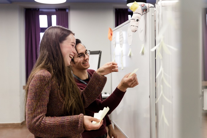 Two young people are standing in front of a white pinboard and seem to be discussing something.