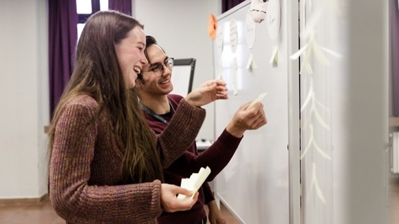 Two young people are standing in front of a white pinboard and seem to be discussing something.