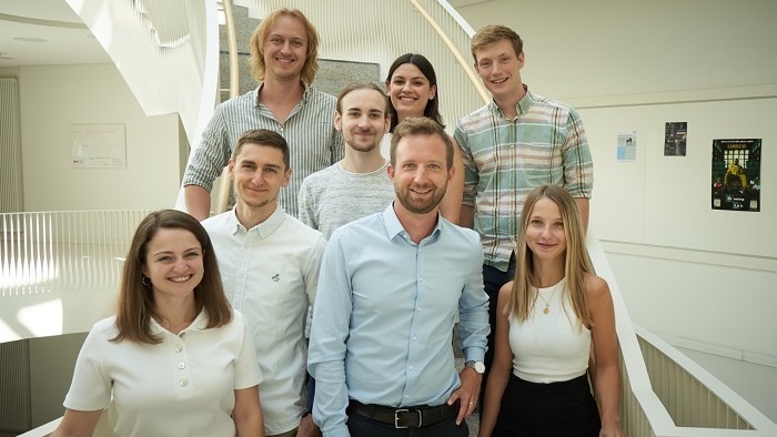 Eight young men and women pose for a group photo in a bright staircase and smile at the camera.