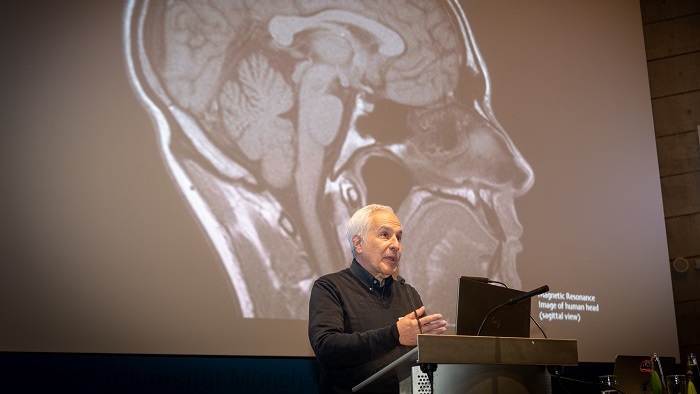 A man dressed in black with white hair stands at the lectern. Behind him is a projection of the human brain.