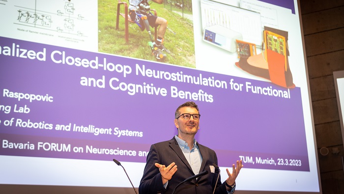 A man with glasses speaks to the audience from the lectern. Behind him, a projection shows another man with a prosthetic leg.