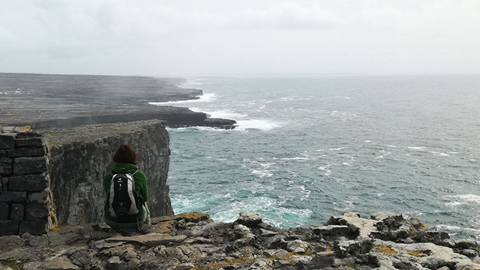 A young woman sits on the edge of a cliff and looks out over the sea. She is sitting with her back to the photographer, who is not in the picture.