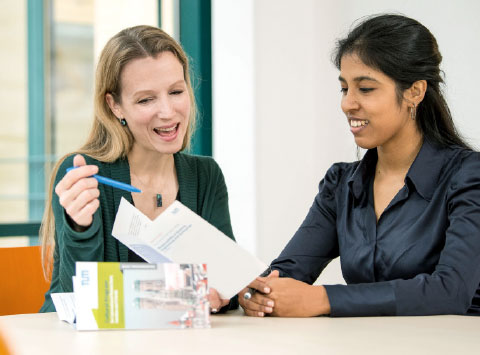 A woman gives advice to another, younger woman. Both of them are sitting at a table.