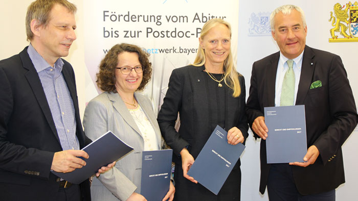 Four formally dressed men and women stand in front of a banner and hold reports in their hands.