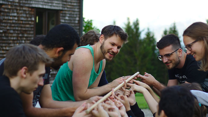 A group of people balance a very thin wooden tube on their fingers.