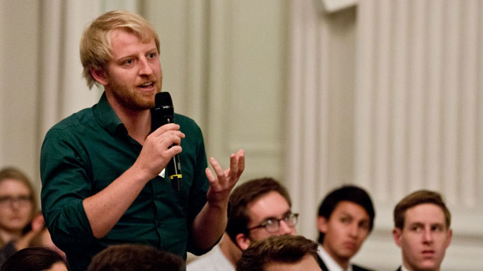 A young man stands in the middle of a seated audience and speaks animatedly to a person not visible in the photo.