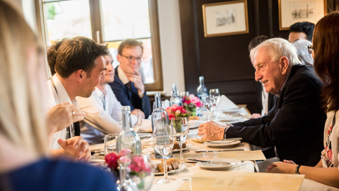 Mehrere junge Menschen unterhalten sich bei einem Abendessen angeregt mit einem älteren Herrn.