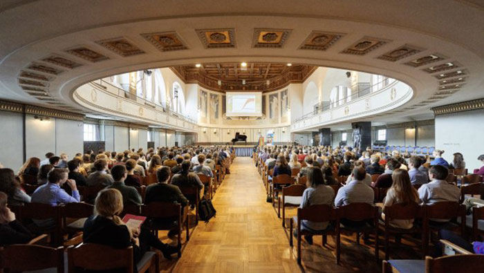 In einem großen Auditorium sitzen viele Menschen und warten auf den Beginn der Veranstaltung.