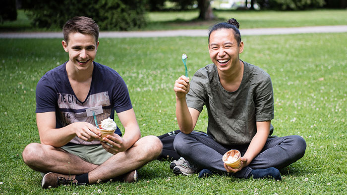 Zwei junge Männer sitzen im Schneidersitz auf einer Wiese und essen Eis aus einem Becher.