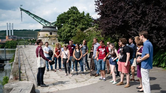 Das Bild zeigt eine Situation, wie man sie aus Stadtführungen kennt: Eine Gruppe junger Menschen hört einem Mann zu, der etwas erläutert. 