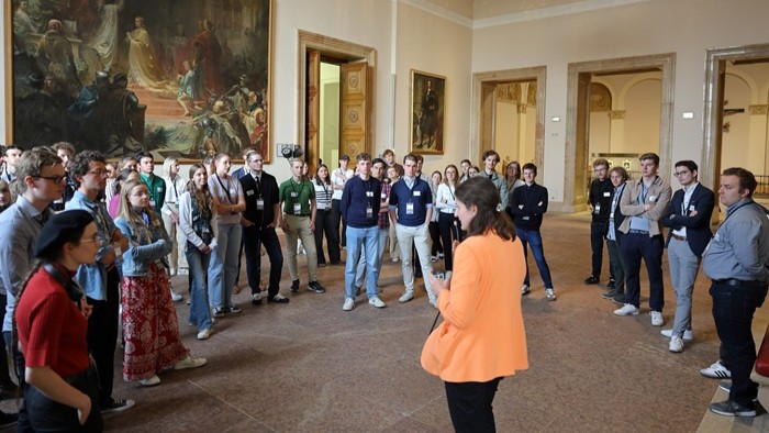 Eine Person in orangefarbener Jacke steht vor einer Gruppe von Menschen in einem großen, historisch ausgestatteten Saal mit Wandgemälden und Kronleuchter.