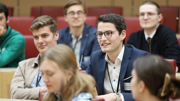 Mehrere junge Personen sitzen in roten Sitzreihen eines Sitzungssaals; eine Person in dunklem Sakko spricht in ein Tischmikrofon.