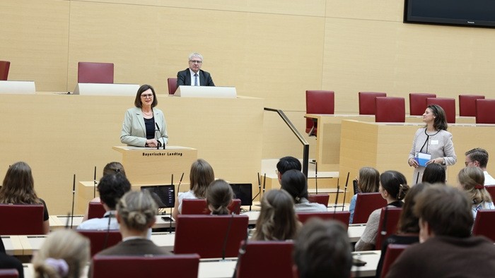 Eine Frau am Rednerpult des Bayerischen Landtags spricht vor einem Publikum in roten Sitzreihen.