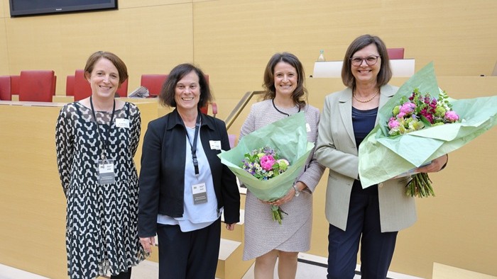 Vier Personen mit Namensschildern stehen in einem hellen Sitzungssaal; zwei von ihnen halten jeweils einen bunten Blumenstrauß.