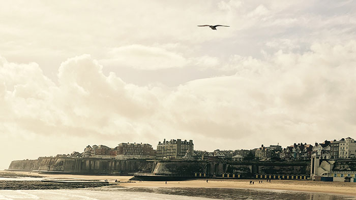 Landschaftsbild: Von einem Kai mit einem hölzernen Geländer aus ist eine Stadtsilhouette mit Strand zu sehen.