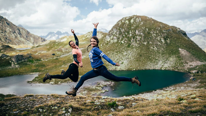 Two young women leap into the air in front of mountainous scenery.