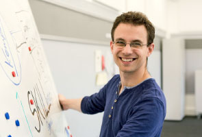 A young man stands in front of a flipchart and smiles at the camera.