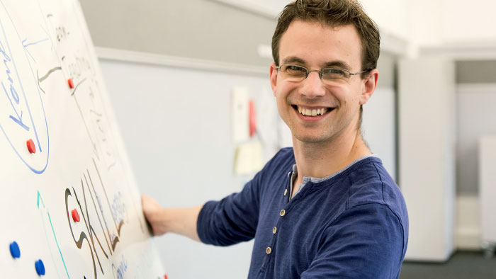 A young man stands in front of a flipchart and smiles at the camera.