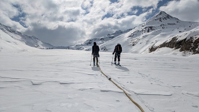 © Céline Walker Two people are climbing up the glacier on touring skis, one of them pulling a yellow tube with two white, bottle-shaped elements behind them. They are surrounded by mountains and there is snow everywhere.